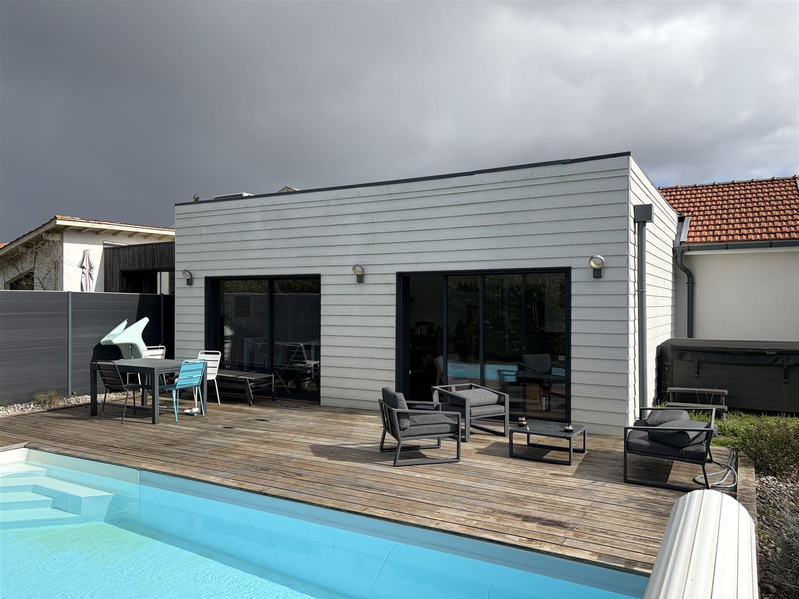 Avant : Terrasse en bois nue équipée d'une pergola bioclimatique anthracite à lames orientables et store latéral, sur la photo réelle du client